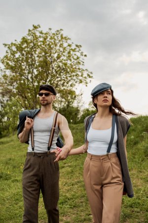 Stylish romantic couple in newsboy caps, suspenders and vintage outfits holding hands while standing with cloudy sky and green field at background, trendy twosome in rustic setting, romantic getaway