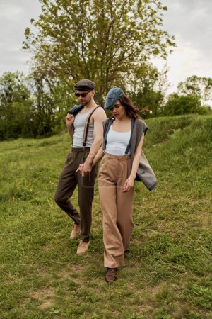 Full length of trendy romantic couple in rural outfits, newsboy caps and suspenders holding hands and walking together on grassy field at summer, trendy twosome in rustic setting