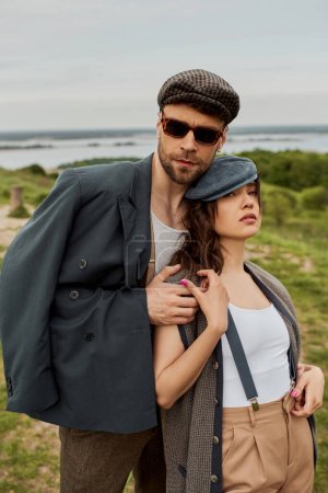 Fashionable man in sunglasses and jacket hugging brunette girlfriend in newsboy cap and suspenders while standing with blurred scenic landscape and sky at background, trendy twosome in rustic setting