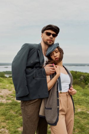 Fashionable bearded man in sunglasses and jacket hugging brunette girlfriend in newsboy cap and suspenders while spending time in rural setting, trendy twosome in rustic setting, romantic getaway