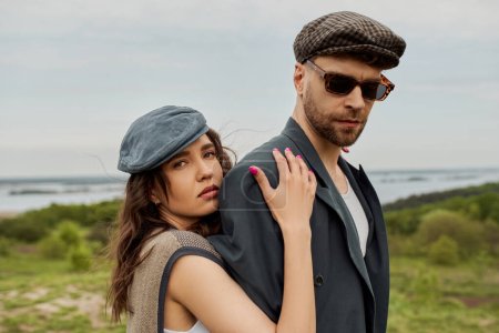 Portrait of trendy brunette woman in newsboy cap and vest hugging boyfriend in sunglasses and jacket while looking at camera with landscape at background, trendy twosome in rustic setting