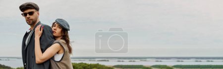 Stylowa brunetka w kamizelce i vintage strój obejmujący chłopaka w kurtce i okularach przeciwsłonecznych patrząc na aparat fotograficzny z krajobrazem w tle, modne dwa w rustykalnym otoczeniu, baner 