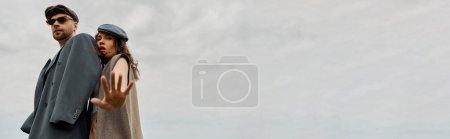 Foto de Vista de ángulo bajo de la mujer de moda en traje vintage mirando a la cámara mientras está de pie cerca de novio barbudo con chaqueta y gafas de sol con cielo nublado en el fondo, pancarta, escapada romántica - Imagen libre de derechos