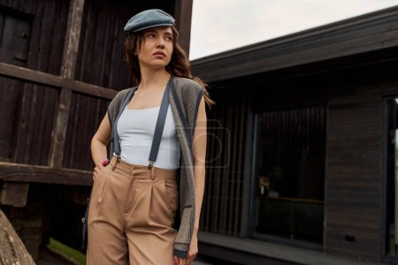 Fashionable brunette woman in vintage outfit, newsboy cap and suspenders holding hand in pocket of pants while standing near rustic house outdoors, vintage-inspired clothing
