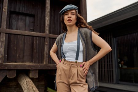 Photo for Stylish brunette woman in vintage outfit and newsboy cap posing in vest and suspenders while standing near rustic house in rural setting at summer, vintage-inspired clothing - Royalty Free Image