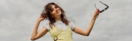 Photo for Low angle view of positive brunette woman in stylish sundress touching hair, looking at camera and holding sunglasses while standing with cloudy sky at background, summertime joy, banner - Royalty Free Image