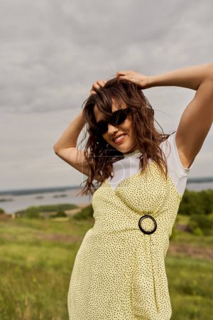 Portrait of joyful and stylish brunette woman in sundress and sunglasses touching head and standing with blurred scenic landscape and sky at background, summertime joy
