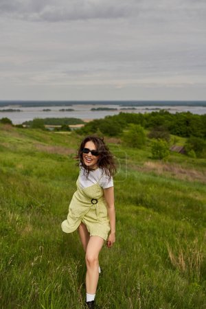 Mujer morena alegre y elegante en gafas de sol y vestido de pie en el prado verde con hierba y pasar tiempo con el paisaje borroso y el cielo en el fondo, alegría de verano