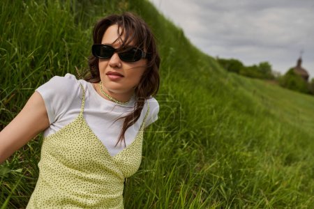 Portrait of trendy brunette woman in sunglasses and stylish sundress sitting on hill with blurred green grass and cloudy sky at background, natural landscape concept