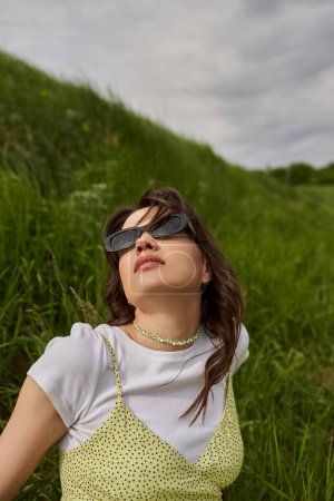 Porträt einer trendigen brünetten Frau mit Sonnenbrille und stylischem Kleid, die auf verschwommenen grasbewachsenen Hügeln sitzt und relaxt, mit verschwommener Landschaft und Himmel im Hintergrund, natürliche Landschaft
