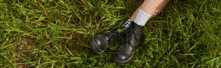 Top view of legs of woman in stylish boots sitting on grassy meadow outdoors, natural landscape
