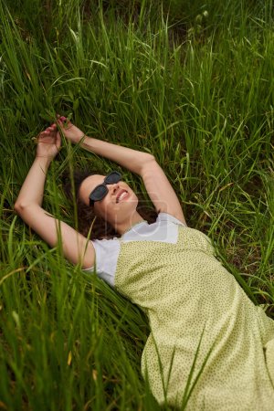 Vista superior de mujer morena alegre y elegante en gafas de sol y vestido de sol tumbado y relajante en el césped cubierto de hierba en verano, paisaje natural y relajante en concepto de naturaleza, paisaje rural