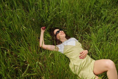 Vue en angle élevé de femme brune positive et élégante en lunettes de soleil et robe de soleil couchée et relaxante sur la prairie avec herbe verte, paysage naturel et concept de détente dans la nature