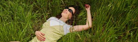 High angle view of cheerful brunette woman in stylish sundress and sunglasses relaxing and lying on meadow with green grass, natural landscape and relaxing in nature concept, banner 
