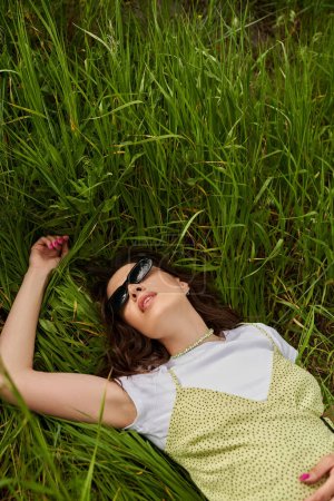 Téléchargez les photos : Vue du dessus de la femme brune à la mode en lunettes de soleil et robe de soleil couché sur l'herbe verte sur le terrain en été, paysage naturel et relaxant dans le concept de la nature, paysage rural - en image libre de droit