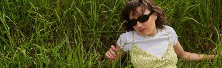 High angle view of stylish brunette woman in sunglasses and sundress lying on meadow and touching green grass, peaceful retreat and relaxing in nature concept, banner, rural landscape