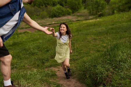 Cheerful brunette woman in stylish sundress and boots holding hand of boyfriend while walking on grassy hill at summer, rural tranquility and relaxing in nature concept