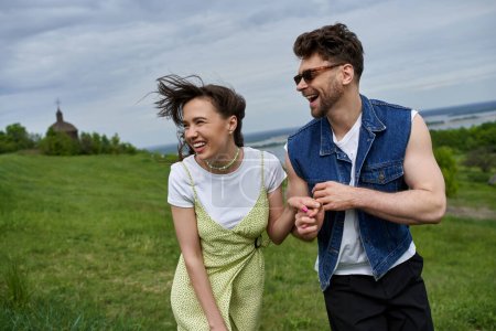 Cheerful bearded man in sunglasses and denim vest holding hand of brunette girlfriend in sundress while walking on grassy field, couple in love enjoying nature and relaxing concept