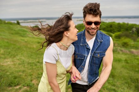 Photo for Cheerful brunette woman in sundress holding hand of stylish boyfriend in sunglasses and denim vest and walking on blurred grassy field, couple in love enjoying nature, tranquility - Royalty Free Image