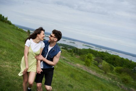 Fröhliches romantisches Paar in stylischem Sommer-Outfit, das Spaß hat und sich beim Spazierengehen auf verschwommenem Grashügel mit Himmel im Hintergrund anschaut, verliebtes Paar, das die Natur genießt, Ruhe