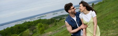 Photo for Cheerful bearded man in sunglasses and denim vest hugging stylish brunette girlfriend in sundress while walking together on blurred grassy field, couple in love enjoying nature, banner - Royalty Free Image