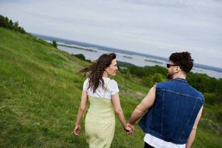 Photo for Side view of smiling and trendy romantic couple in summer outfits holding hands while walking on grassy hills with cloudy sky at background, couple in love enjoying nature, tranquility - Royalty Free Image