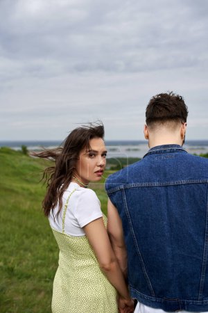 Photo for Fashionable brunette woman in summer sundress looking at camera and holding hand of boyfriend in denim vest and standing on blurred green field, couple in love enjoying nature, tranquility - Royalty Free Image