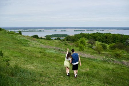Rückansicht des stilvollen Paares im Sommer-Outfit Händchen haltend und stehend auf einer Wiese mit malerischer Aussicht und bewölktem Himmel im Hintergrund, verliebtes Paar genießt die Natur, Ruhe