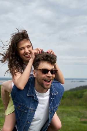 Photo for Cheerful brunette woman piggybacking on stylish boyfriend in sunglasses and denim vest while standing with blurred landscape at background, countryside adventure and love story, tranquility - Royalty Free Image
