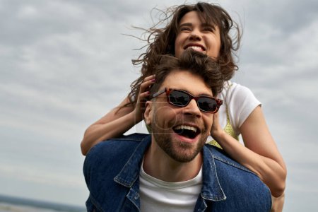 Cheerful brunette woman piggybacking on stylish boyfriend in sunglasses and denim vest and having fun together with cloudy sky at background, countryside adventure and love story
