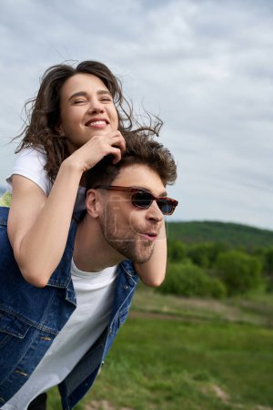 Happy brunette woman piggybacking on excited boyfriend in sunglasses and denim vest and having fun together with blurred nature at background, countryside adventure and love story
