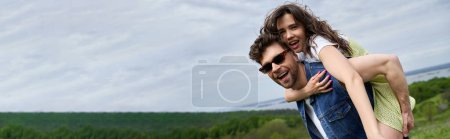 Cheerful brunette woman in trendy sundress piggybacking on bearded boyfriend in sunglasses and denim vest and spending time on nature, countryside adventure and love story, banner 