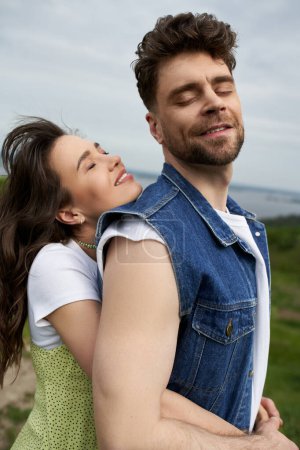 Portrait of brunette woman in sundress smiling and hugging bearded boyfriend in denim vest and closing eyes while standing with nature at background, love story and countryside adventure