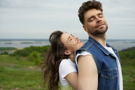 Cheerful brunette woman with closed eyes standing next to stylish bearded boyfriend in denim vest and spending time together with landscape at background, love story and countryside adventure