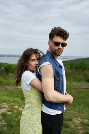 Stylish woman in sundress embracing bearded boyfriend in sunglasses and denim vest and looking at camera while standing with landscape and sky at background, love story and countryside adventure