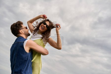 Cheerful bearded man in denim vest lifting brunette girlfriend in summer outfit and sunglasses and standing with cloudy sky at background, love story and countryside adventure, tranquility