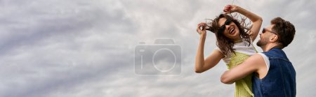 Smiling bearded man in denim vest lifting and hugging cheerful stylish girlfriend in sundress and sunglasses while standing with cloudy sky at background, love story and countryside adventure, banner