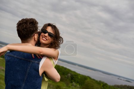 Photo for Smiling brunette woman in summer outfit and sunglasses embracing stylish boyfriend in denim vest with rural landscape and cloudy sky at background, love story and countryside adventure - Royalty Free Image