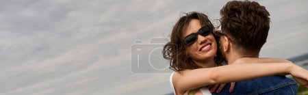 Femme brune positive en lunettes de soleil embrassant petit ami en gilet en denim élégant tout en passant du temps à l'extérieur avec ciel nuageux en arrière-plan, histoire d'amour et aventure à la campagne, bannière