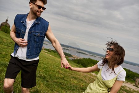 Smiling bearded man in sunglasses and stylish denim vest holding hand of cheerful girlfriend in sundress and spending time on grassy hill at background, countryside wanderlust and love concept