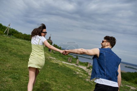Photo for Side view of positive romantic couple in sunglasses and summer outfits holding hands while running on grassy hill with blurred landscape at background, countryside wanderlust and love concept - Royalty Free Image