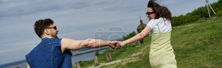 Photo for Side view of cheerful brunette woman in sunglasses and stylish sundress holding hand of bearded boyfriend while running on grassy hill, countryside wanderlust and love concept, banner - Royalty Free Image