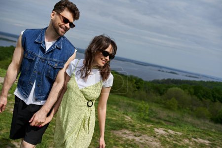 Positive romantic couple in sunglasses and trendy summer outfits holding hands and looking away while walking on blurred landscape, countryside wanderlust and love concept, tranquility