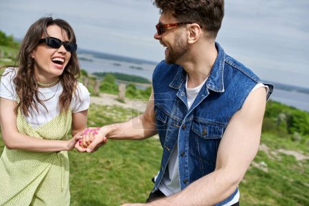 Cheerful brunette woman in trendy summer outfit and sunglasses holding hand of boyfriend in denim vest while spending time in blurred rural setting at background, countryside leisurely stroll
