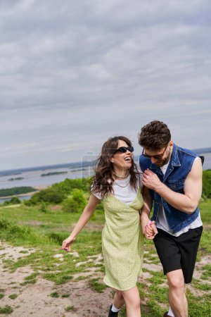 Cheerful brunette man in stylish summer outfit and denim vest holding hand of excited girlfriend in sunglasses and sundress and walking together on path on grassy hill, countryside leisurely stroll