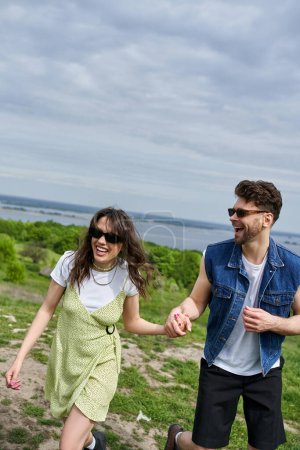 Positive brunette man in sunglasses and denim vest holding hand of cheerful girlfriend in stylish sundress and spending time with nature at background, countryside leisurely stroll, tranquility