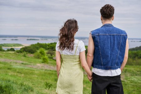 Back view of stylish romantic couple in summer outfits holding hands while standing together on grassy hill with blurred scenic landscape at background, countryside retreat concept