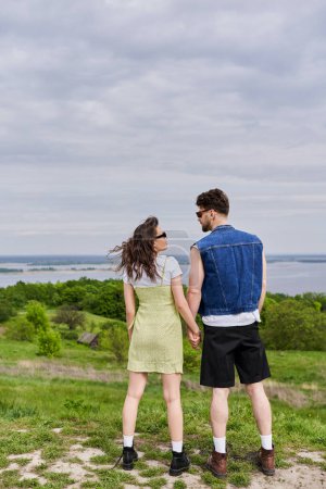 Photo for Full length of stylish brunette romantic couple in sunglasses, summer outfits and boots holding hands and spending time while standing on hill with grass, countryside retreat concept - Royalty Free Image