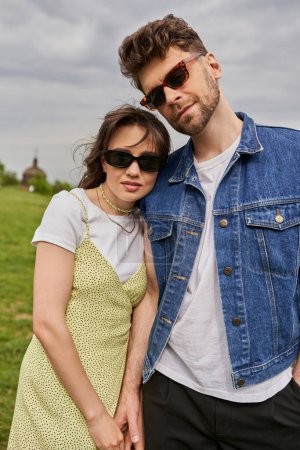 Retrato de pareja romántica de moda en gafas de sol y trajes de verano tocando la mano y posando con paisaje borroso en el fondo, concepto de retiro rural, tranquilidad
