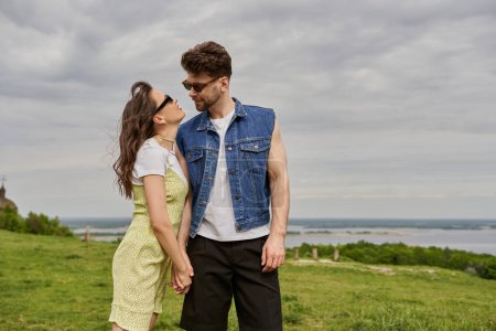 Romantic brunette woman in sunglasses and sundress holding hand of bearded boyfriend in denim vest and standing together with rural landscape and cloudy sky at background, countryside retreat concept